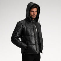 A man wearing a black hooded puffer jacket with quilted stitching, photographed against a plain light gray studio background.