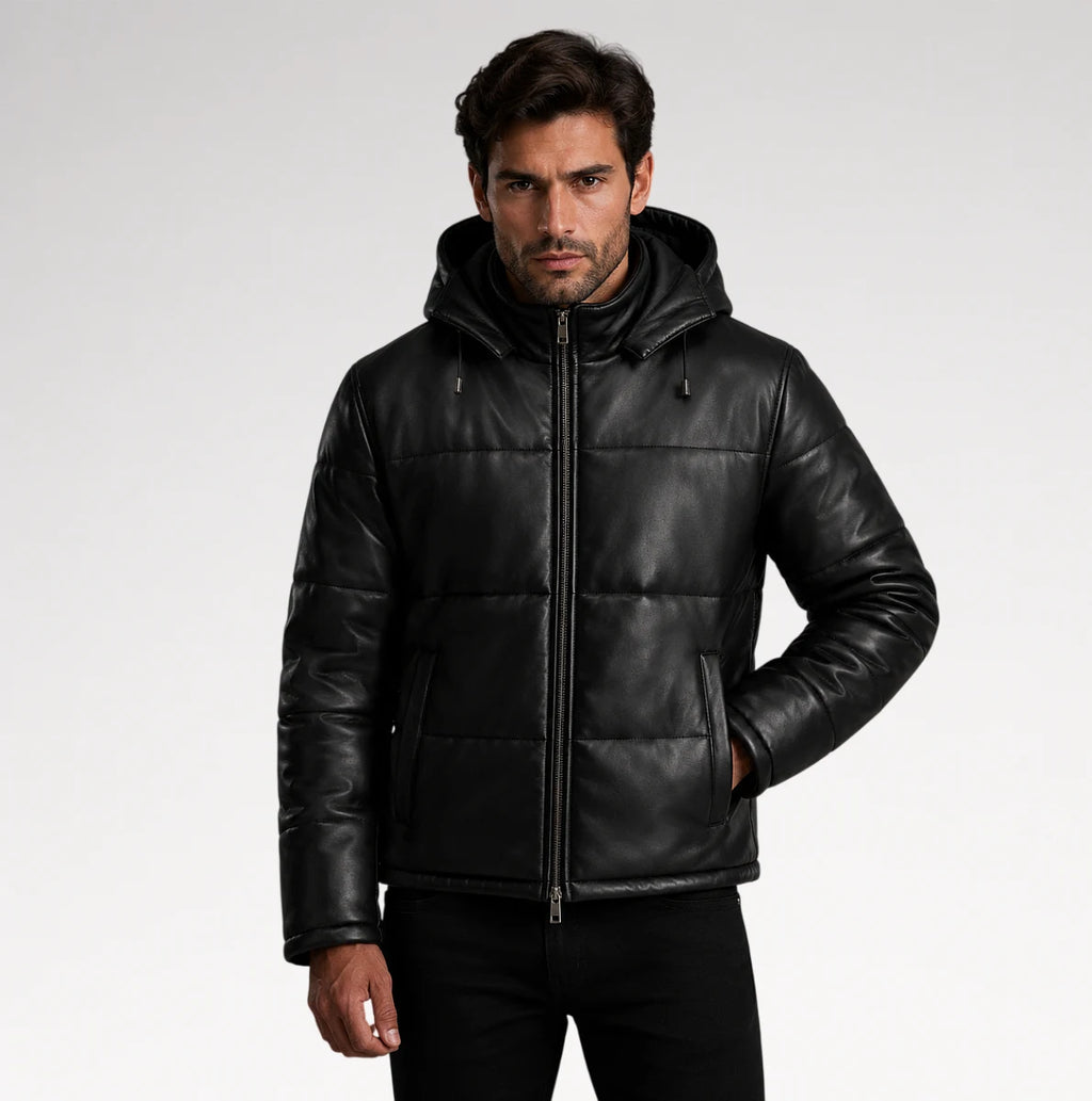 A man wearing a black leather hooded puffer jacket with a front zipper and horizontal quilted stitching, photographed against a light gray studio background.