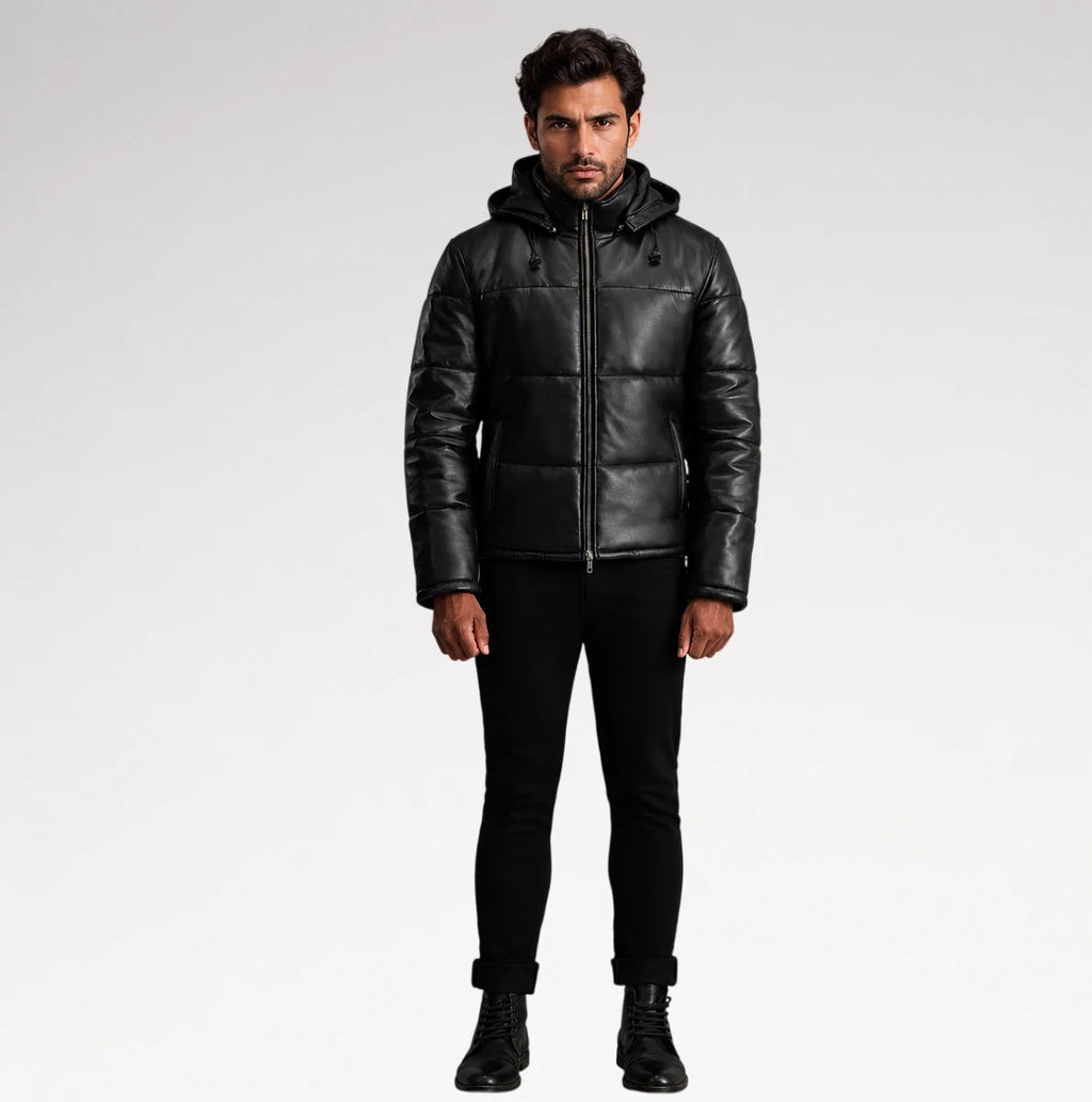 A man modeling a black leather hooded puffer jacket, paired with black pants and boots, against a light gray studio background.