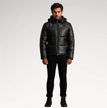 A man modeling a black leather hooded puffer jacket, paired with black pants and boots, against a light gray studio background.