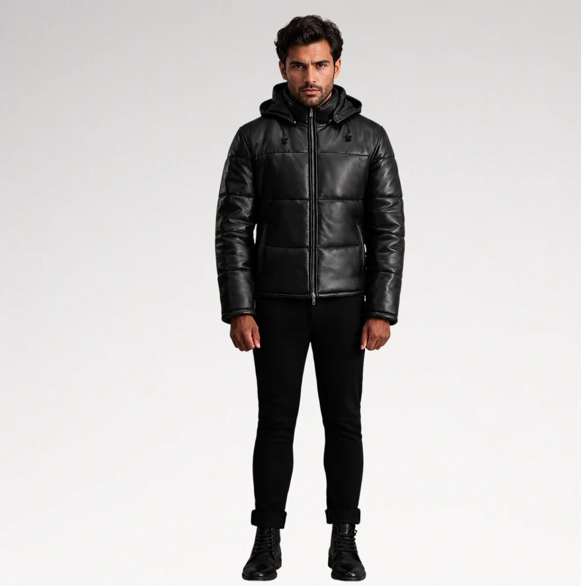 A man modeling a black leather hooded puffer jacket, paired with black pants and boots, against a light gray studio background.
