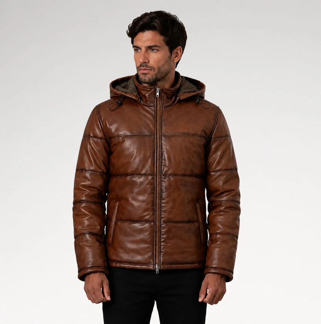 A man wearing a brown leather hooded puffer jacket with horizontal quilting, standing against a light gray studio background.