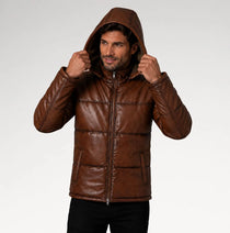 A man wearing a brown faux-leather hooded puffer jacket, holding the hood up. The jacket features horizontal quilting, a full-length front zipper, and zippered hand pockets.