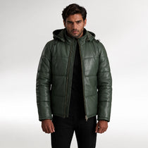 A man wearing a dark green quilted leather hooded jacket over a black shirt and black pants, standing against a light gray studio background.