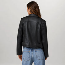 A woman viewed from the back wearing a black leather jacket and light blue jeans against a plain light gray studio background.