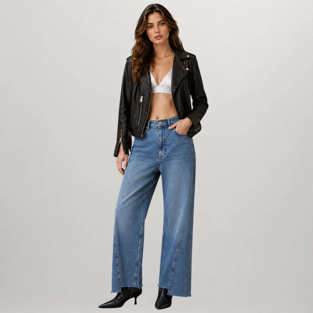 A model wearing a black leather moto jacket, wide-leg blue denim jeans, a white bralette, and black pointed-toe heels, standing against a light gray studio background.