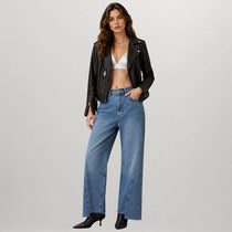 A model wearing a black leather moto jacket, wide-leg blue denim jeans, a white bralette, and black pointed-toe heels, standing against a light gray studio background.