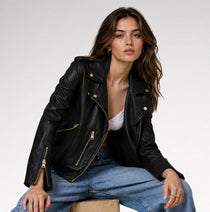 A woman wearing a black leather moto jacket with gold-tone hardware, styled with blue jeans and a white top, sitting against a plain light gray studio background.