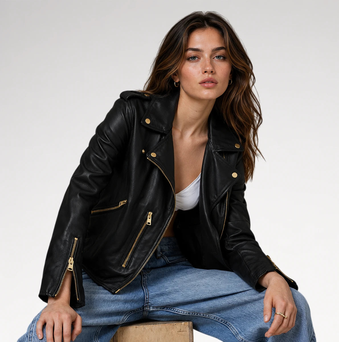 A woman wearing a black leather moto jacket with gold-tone hardware, styled with blue jeans and a white top, sitting against a plain light gray studio background.