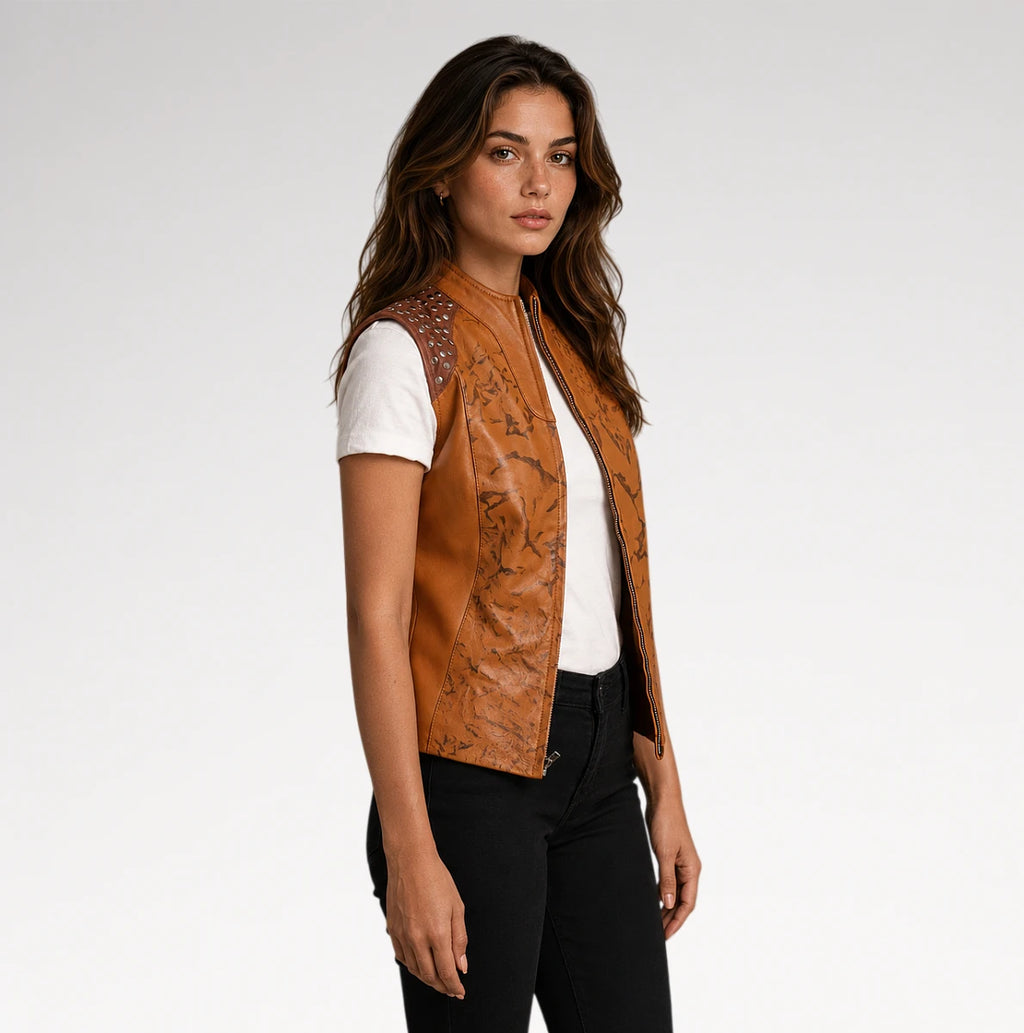 A woman modeling a tan faux-leather vest with black floral print and silver rhinestone shoulder details, worn over a white t-shirt and black pants against a light gray studio background.