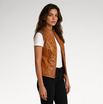 A woman modeling a tan faux-leather vest with black floral print and silver rhinestone shoulder details, worn over a white t-shirt and black pants against a light gray studio background.