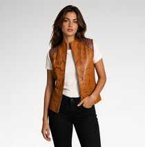 A woman modeling a tan leather vest with dark brown graphic print and silver studs on the shoulders, worn over a white t-shirt and black jeans against a light gray studio background.