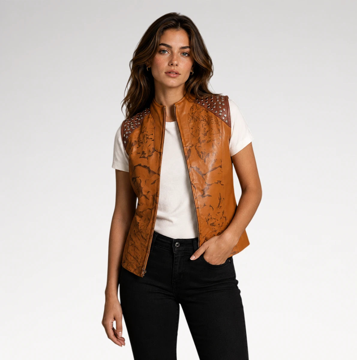 A woman modeling a tan leather vest with dark brown graphic print and silver studs on the shoulders, worn over a white t-shirt and black jeans against a light gray studio background.