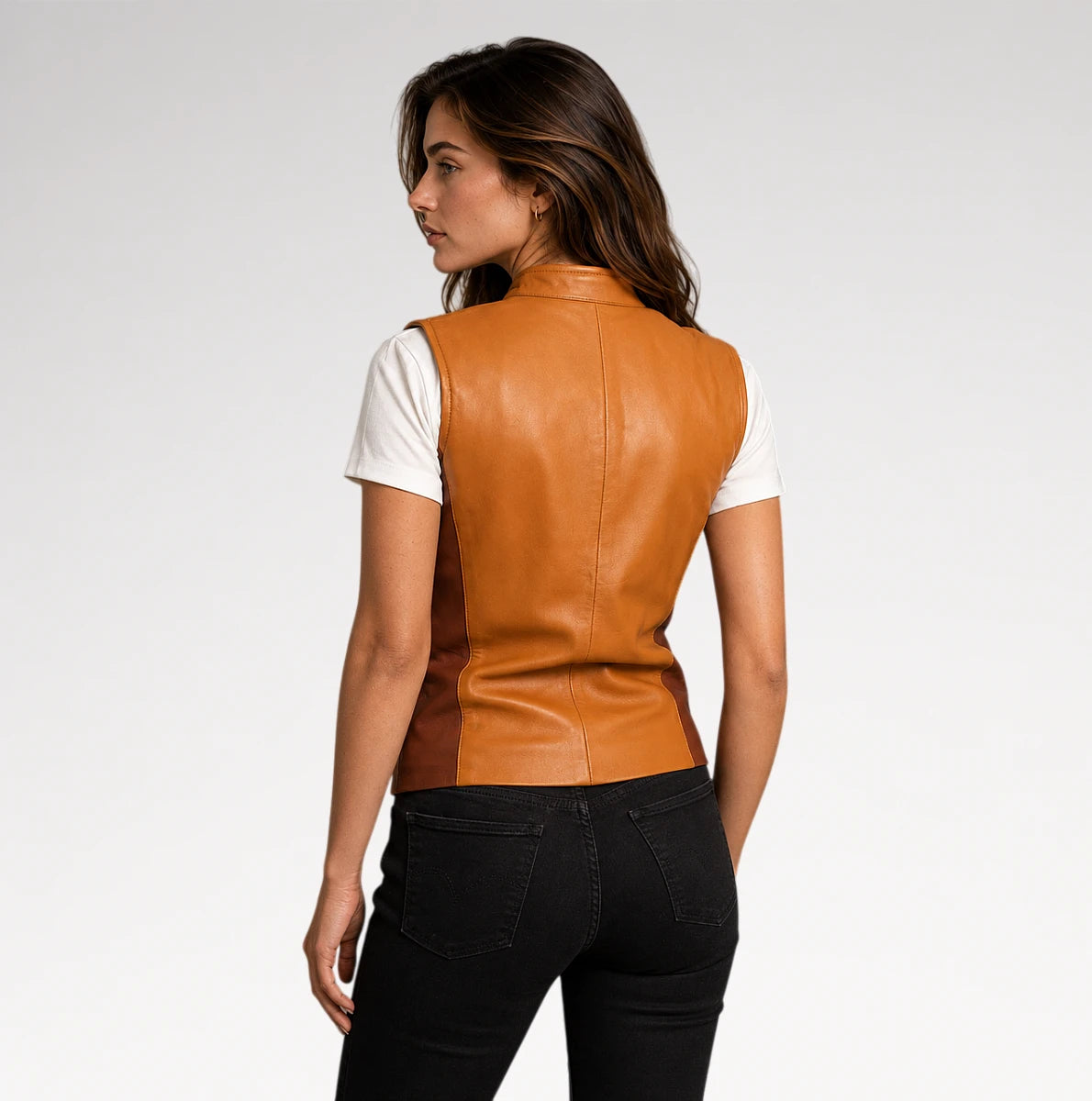 A woman shown from the back wearing a tan leather vest over a white short-sleeve top and black jeans, photographed against a light gray studio background.