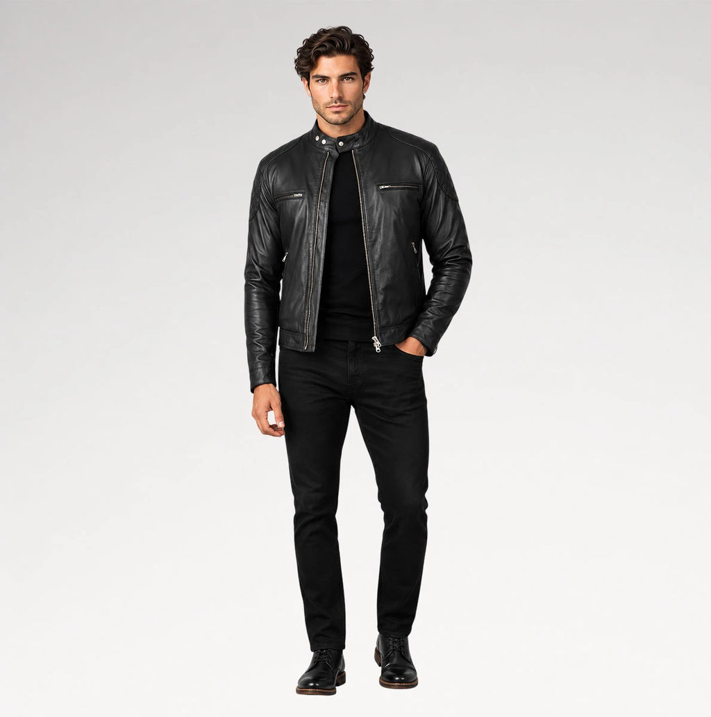 A man modeling a black leather motorcycle jacket over a black shirt and black jeans, standing against a light gray studio background.