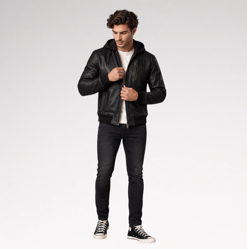 A man modeling a black leather hooded jacket, worn open over a white t-shirt, black skinny jeans, and black low-top sneakers, against a light gray studio background.