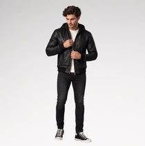 A man modeling a black leather hooded jacket, worn open over a white t-shirt, black skinny jeans, and black low-top sneakers, against a light gray studio background.