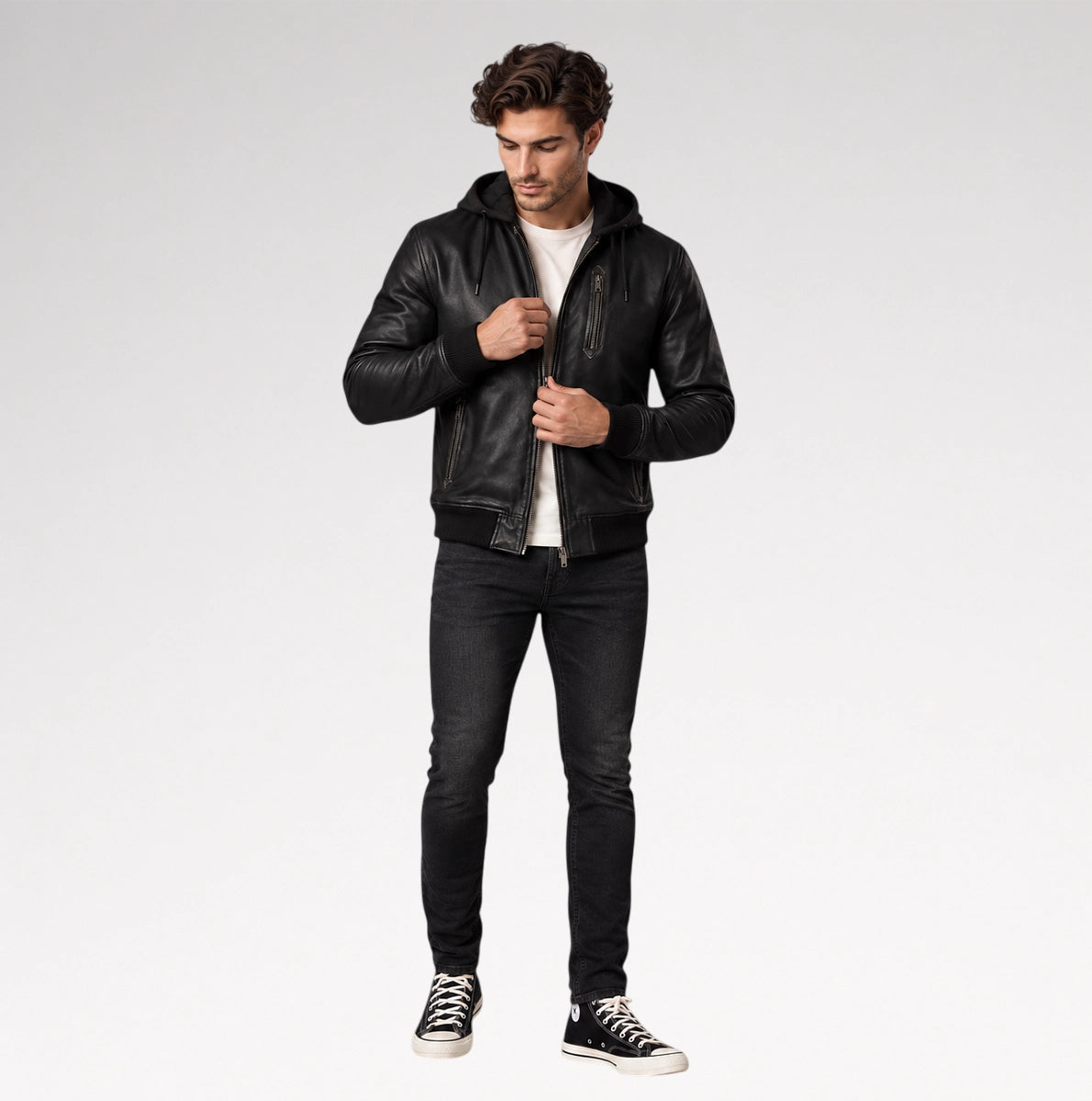 A man modeling a black leather hooded jacket, worn open over a white t-shirt, black skinny jeans, and black low-top sneakers, against a light gray studio background.