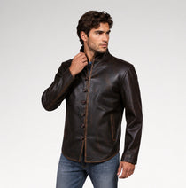 A man wearing a dark brown leather jacket with a mandarin collar and button closure, paired with blue jeans, photographed against a light gray studio background.