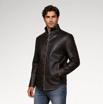 A man wearing a dark brown leather jacket with a high stand-up collar and decorative front buttons, paired with blue jeans, photographed against a light gray studio background.