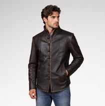 A man wearing a dark brown leather jacket with a mandarin collar and button closure, paired with blue jeans, photographed against a light gray studio background.