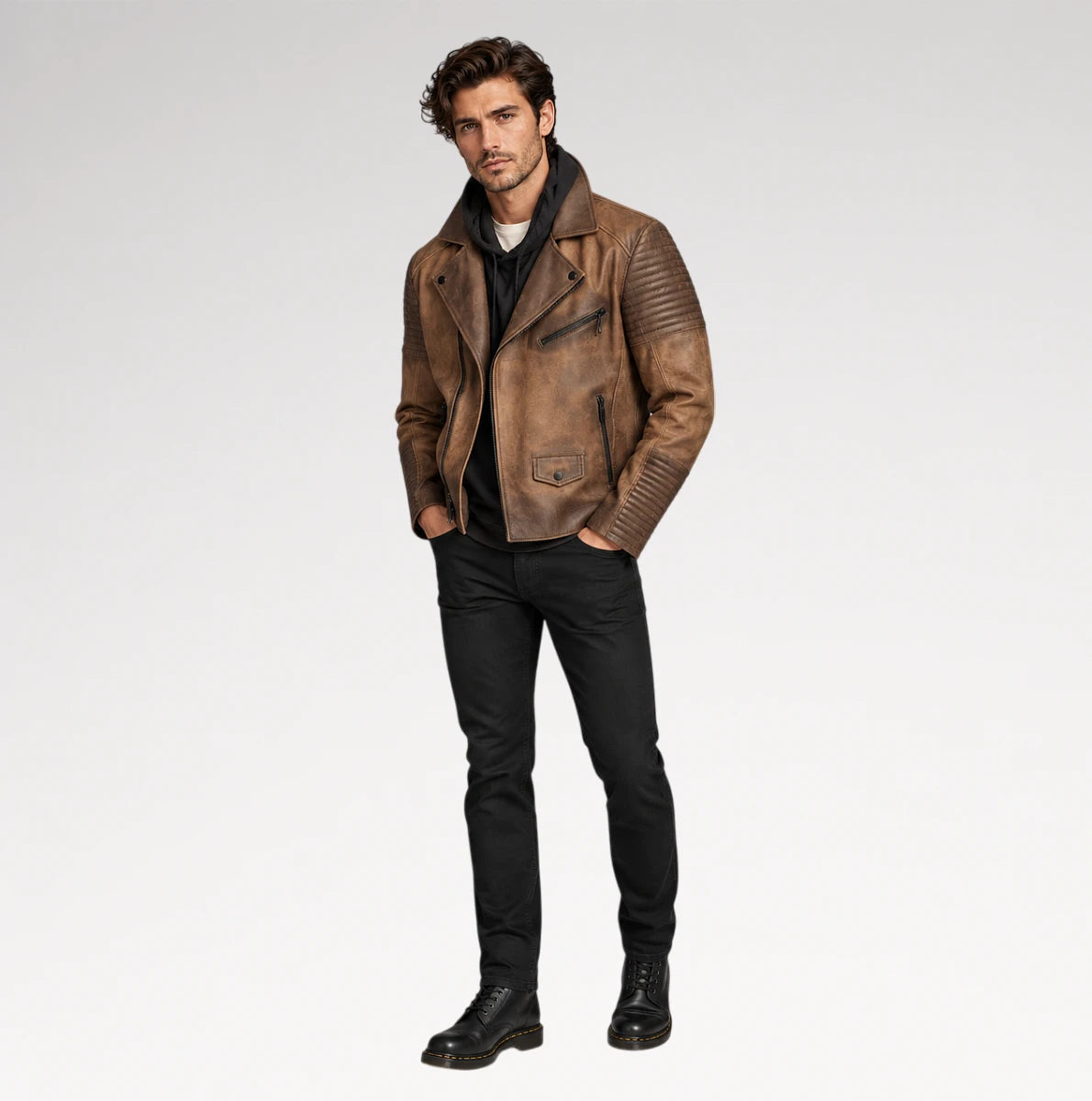 A man modeling a distressed brown leather moto jacket with black pants and black leather boots, standing against a light gray studio background.