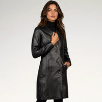 A woman modeling a black faux-leather double-breasted coat over a black turtleneck, photographed against a light gray studio background.