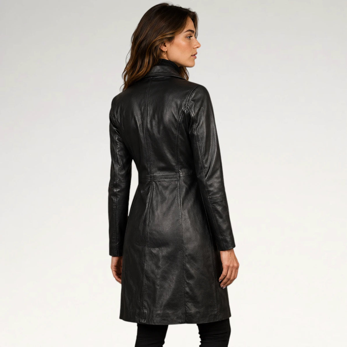 A woman wearing a black leather trench coat, shown from the back and side profile against a plain white studio background.