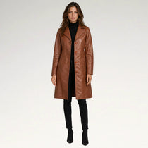 A woman modeling a mid-length brown faux-leather coat over a black turtleneck, black pants, and black ankle boots, photographed against a seamless light gray studio background.
