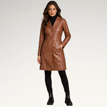 A woman modeling a brown faux-leather coat over a black turtleneck, black leggings, and black ankle boots, standing against a light gray studio background.