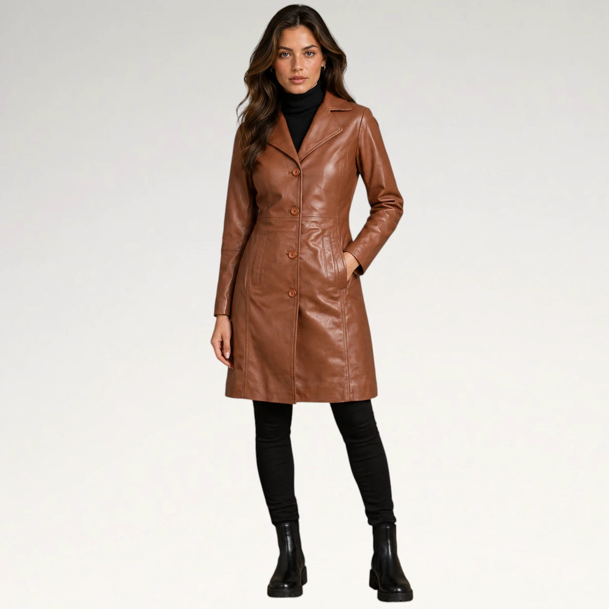 A woman modeling a brown faux-leather coat over a black turtleneck, black leggings, and black ankle boots, standing against a light gray studio background.