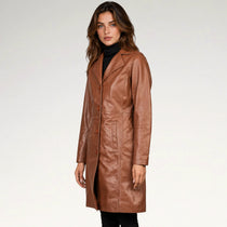 A woman modeling a mid-length, brown faux-leather coat over a black turtleneck, photographed against a clean, light gray studio background.