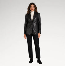 A woman modeling a black leather blazer over a white turtleneck sweater, paired with black trousers and black ankle boots, against a plain light gray studio background.