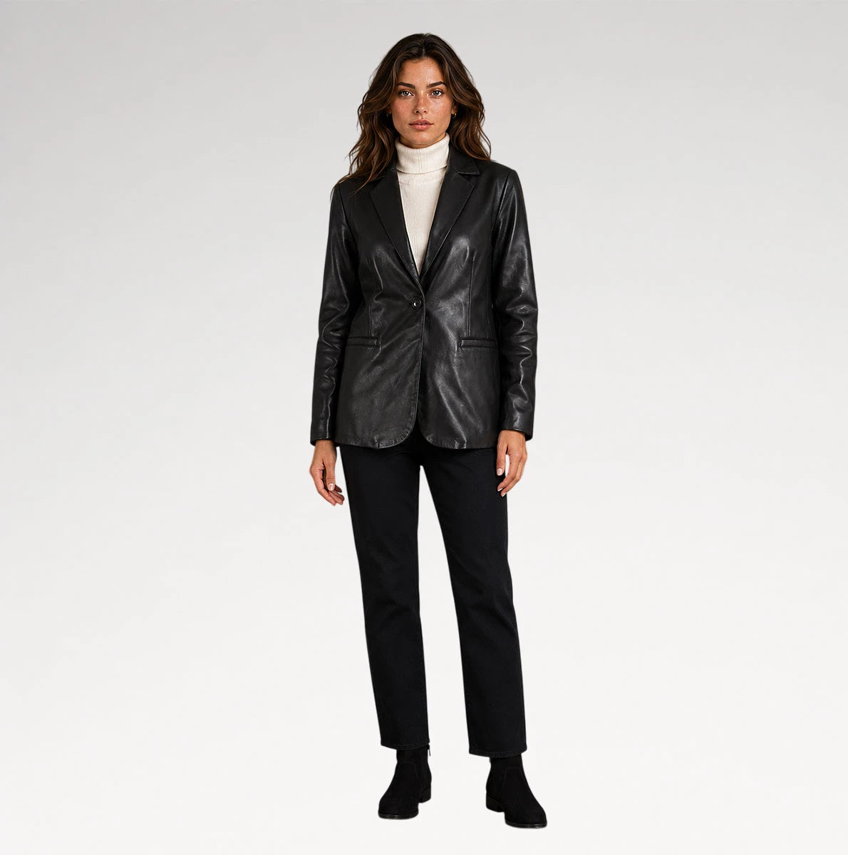 A woman modeling a black leather blazer over a white turtleneck sweater, paired with black trousers and black ankle boots, against a plain light gray studio background.
