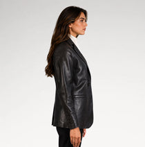 A woman shown in side profile wearing a black leather blazer over a white turtleneck, photographed against a clean light gray studio background.
