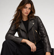A woman wearing a black leather moto jacket with gold-tone zippers, styled with a black ribbed top and black trousers, photographed against a clean light gray studio background.