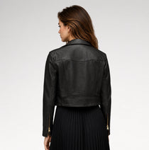 A woman shown from the back wearing a cropped black leather jacket with gold-tone zipper details on the sleeves, paired with a black pleated skirt, against a light gray studio background.