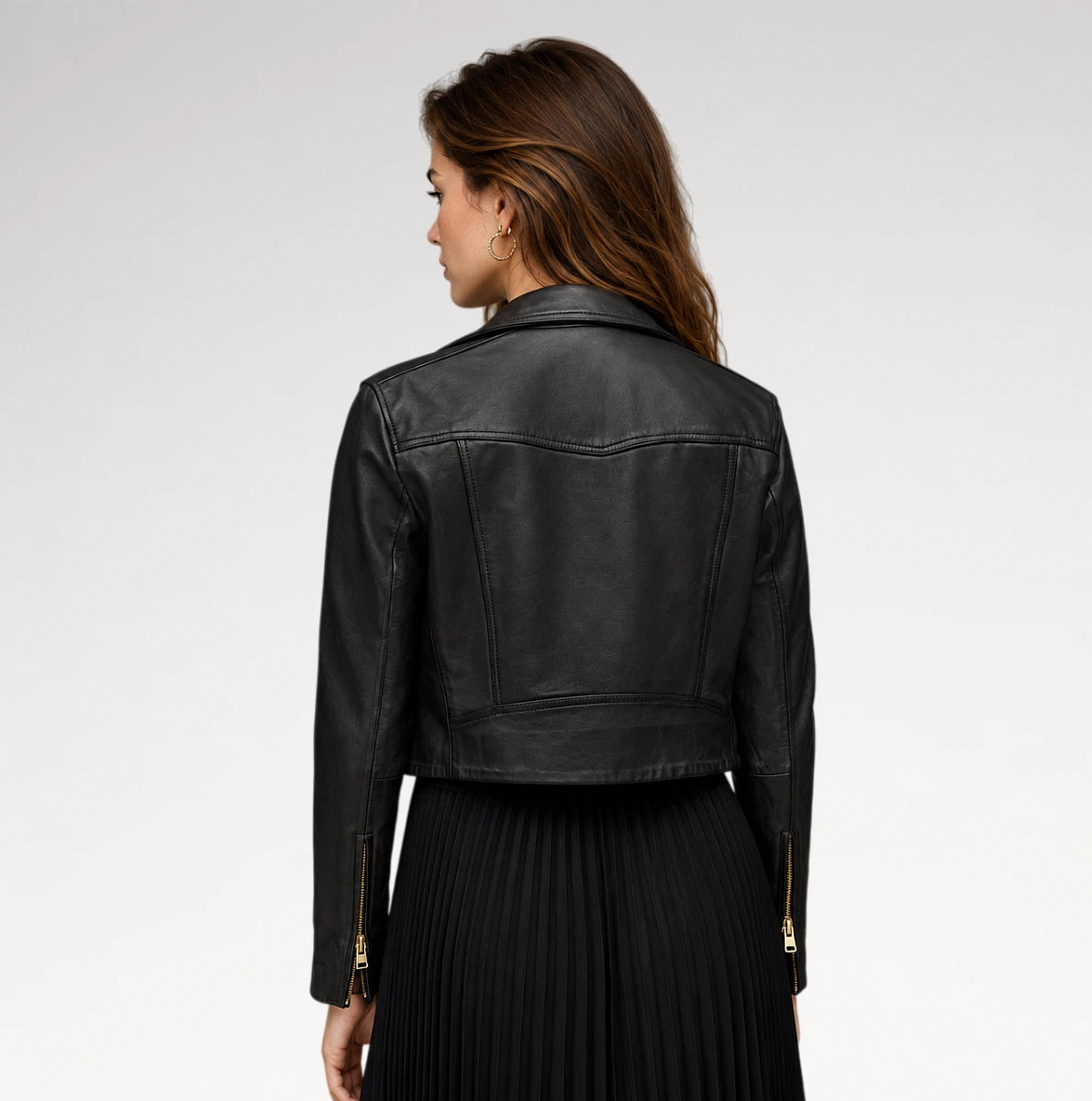 A woman shown from the back wearing a cropped black leather jacket with gold-tone zipper details on the sleeves, paired with a black pleated skirt, against a light gray studio background.