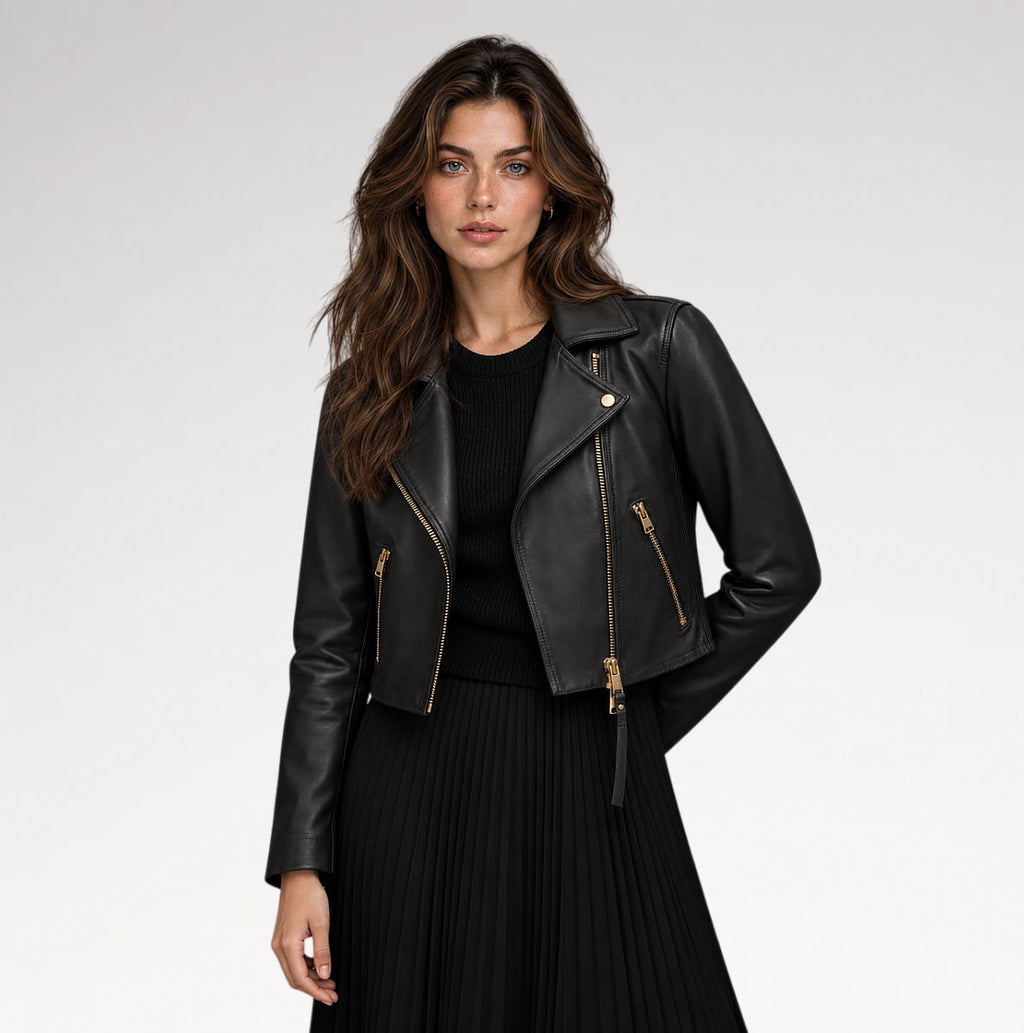A woman modeling a black faux-leather moto jacket with gold-tone zippers, worn over a black ribbed top and black pleated skirt, against a light gray studio background.