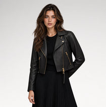 A woman modeling a black faux-leather moto jacket with gold-tone zippers, worn over a black ribbed top and black pleated skirt, against a light gray studio background.