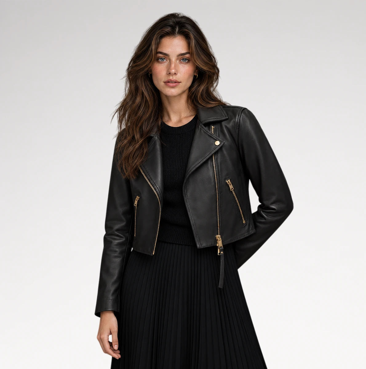 A woman modeling a black faux-leather moto jacket with gold-tone zippers, worn over a black ribbed top and black pleated skirt, against a light gray studio background.