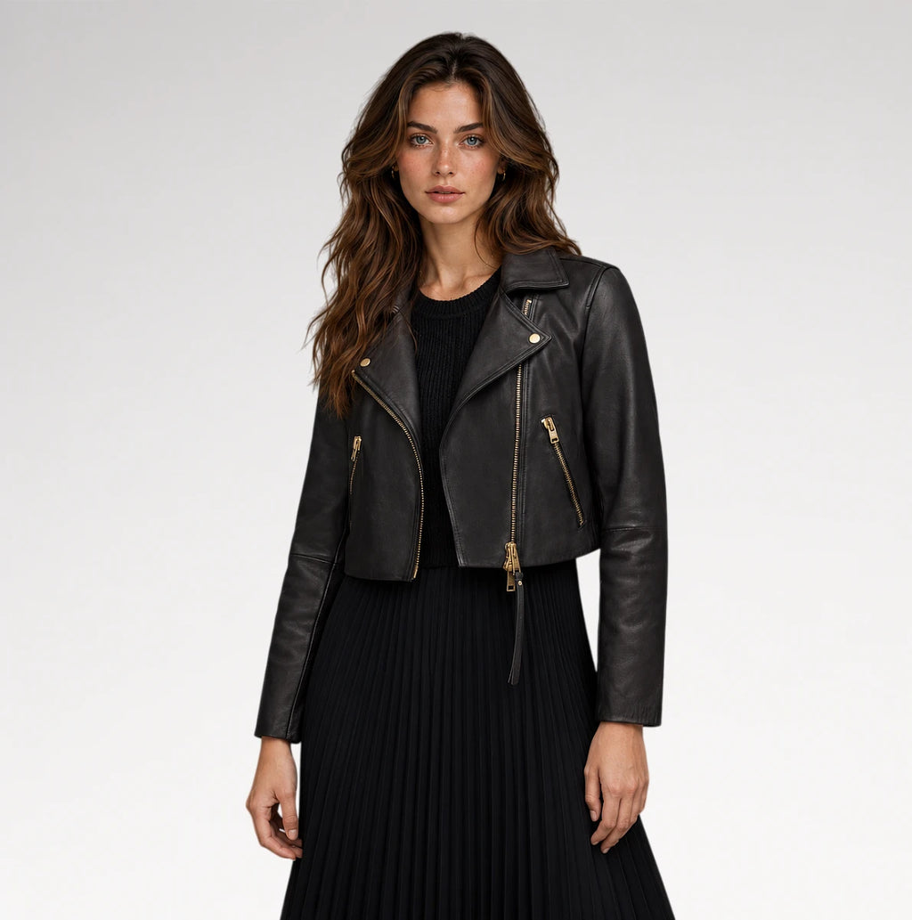 A woman modeling a cropped black leather moto jacket with gold-tone zippers, worn over a black top and black pleated skirt, against a light gray studio background.