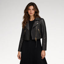 A woman modeling a cropped black leather moto jacket with gold-tone zippers, worn over a black top and black pleated skirt, against a light gray studio background.