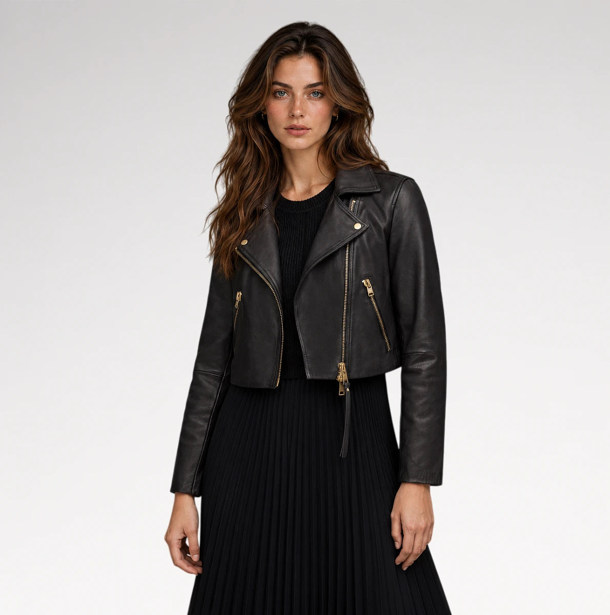 A woman modeling a cropped black leather moto jacket with gold-tone zippers, worn over a black top and black pleated skirt, against a light gray studio background.