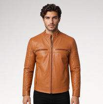A man wearing a tan leather moto jacket with a front zipper and zippered chest pockets, paired with black pants, against a light gray studio background.