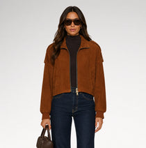 A woman modeling a cropped brown suede jacket over a black turtleneck, paired with dark blue jeans and holding a brown handbag, set against a plain light gray studio background.