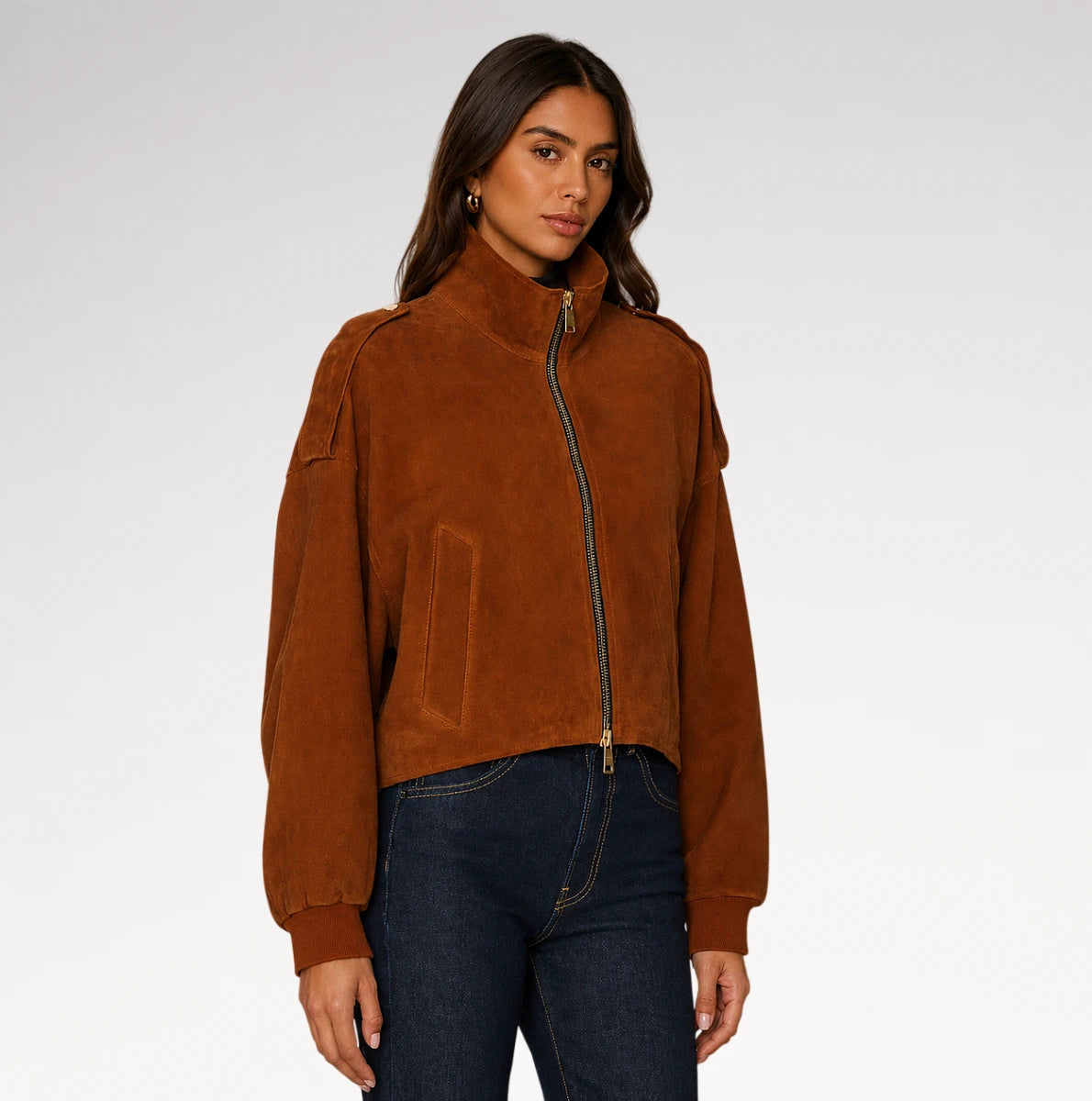 A woman modeling a cropped brown suede jacket with a high collar and front zipper, paired with dark blue jeans, against a light gray studio background.