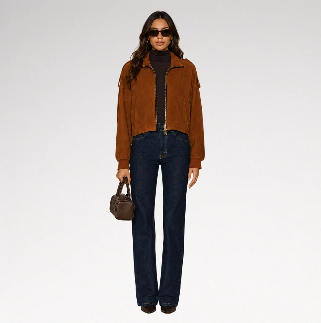 A model stands against a light gray studio background wearing a brown suede cropped jacket over a black turtleneck, dark blue jeans, sunglasses, and holding a small brown handbag.