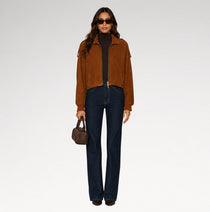 A model stands against a light gray studio background wearing a brown suede cropped jacket over a black turtleneck, dark blue jeans, sunglasses, and holding a small brown handbag.