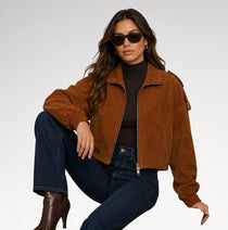 A woman wearing a brown suede bomber jacket, dark brown turtleneck, dark blue jeans, and brown leather boots, posing against a light gray studio background.